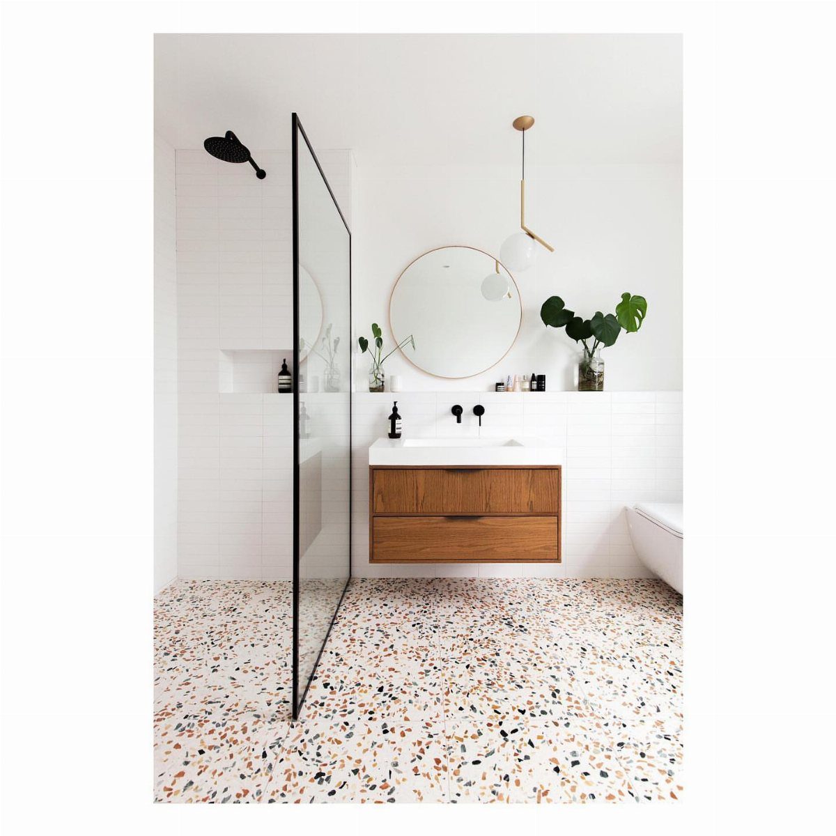 White terrazzo flooring in a bright bathroom with a wooden vanity and round mirror.