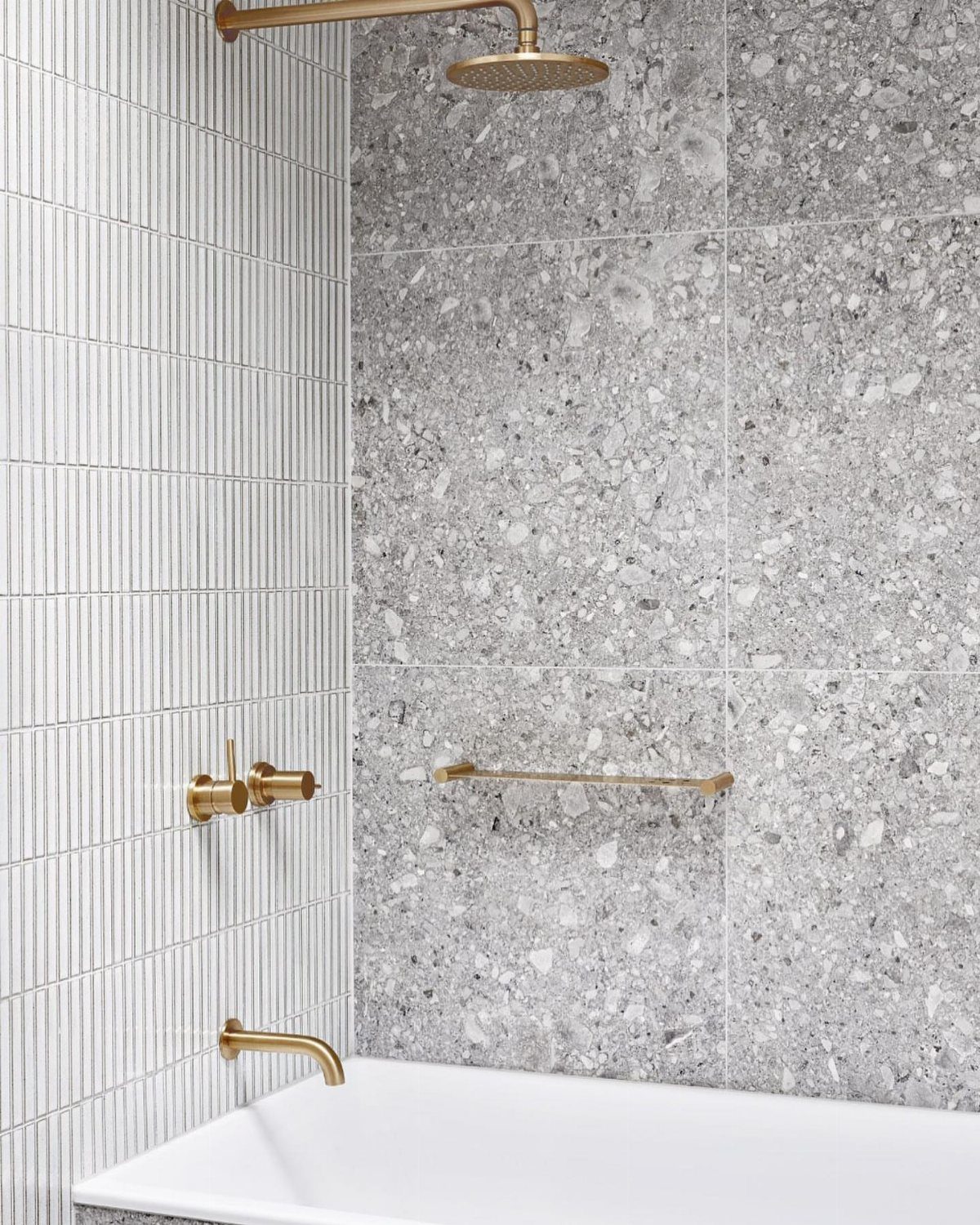 Grey terrazzo tiles on a shower wall with gold fixtures and white bathtub.