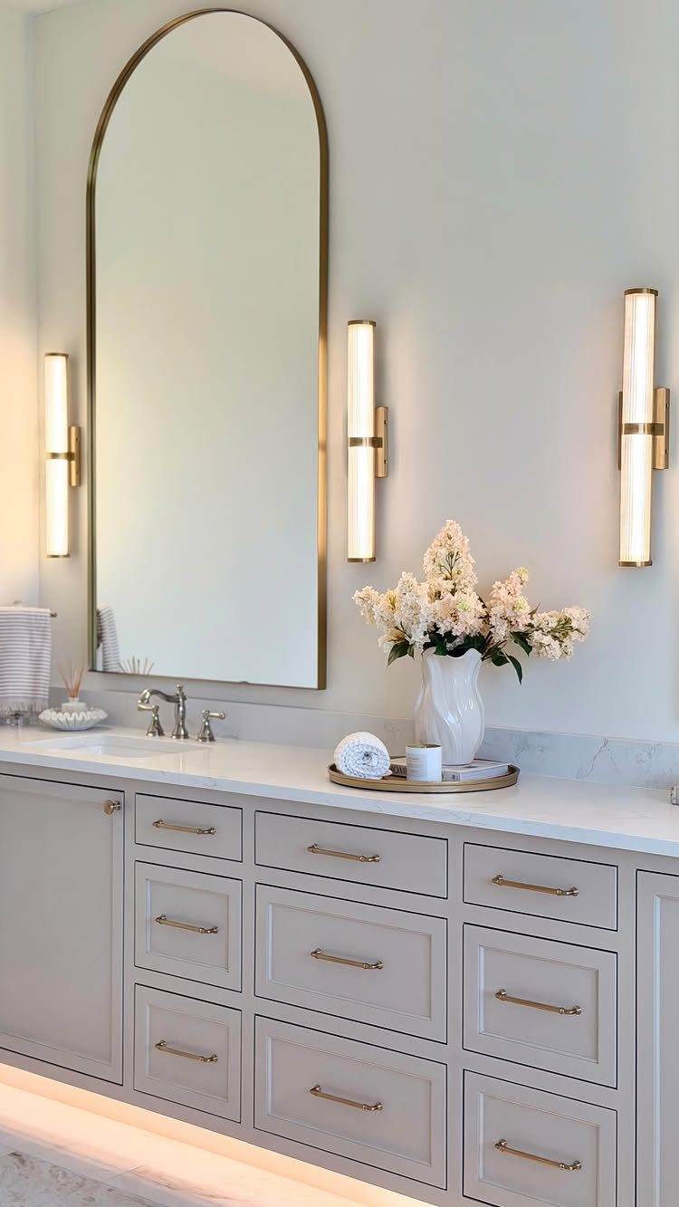 Arched gold-framed mirror above a light gray vanity with gold hardware and white marble.