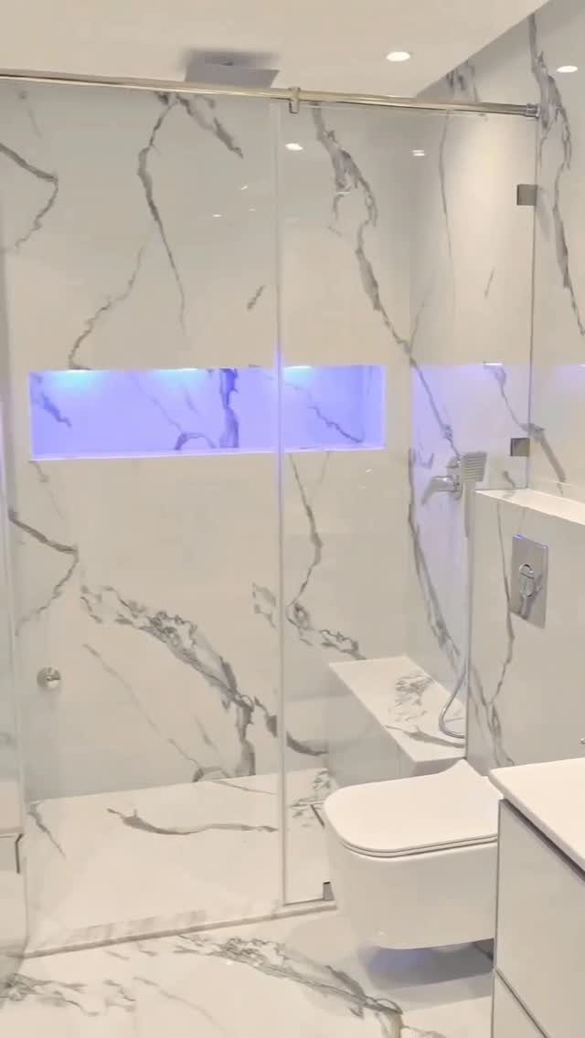 Modern bathroom with white marble walls, glass shower, and blue LED niche lighting.