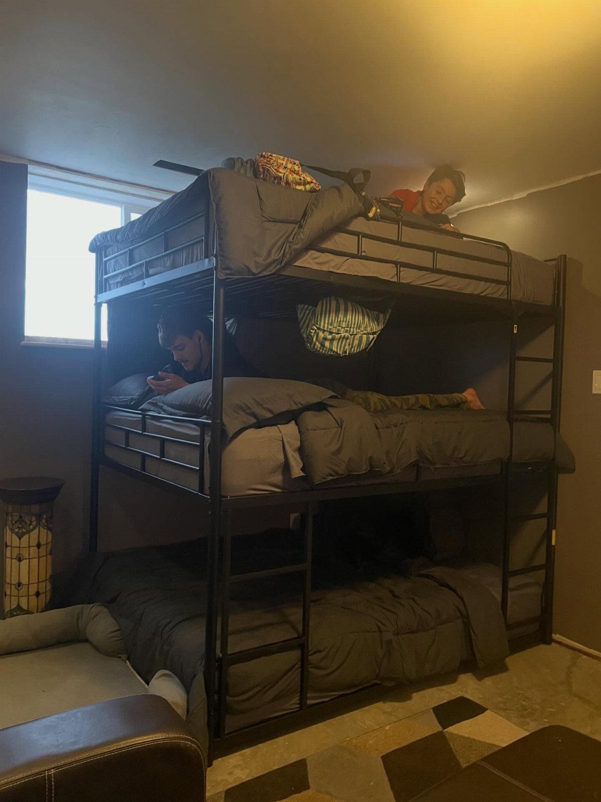 Black metal triple bunk bed with grey bedding and children in each bunk.