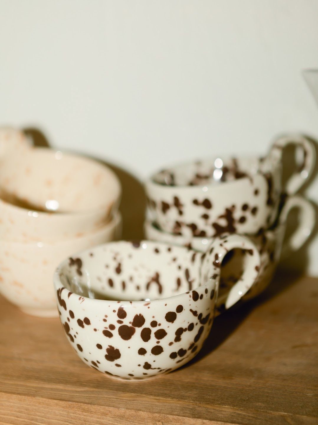 Beige ceramic mugs with brown spotted patterns sitting on a wooden shelf.