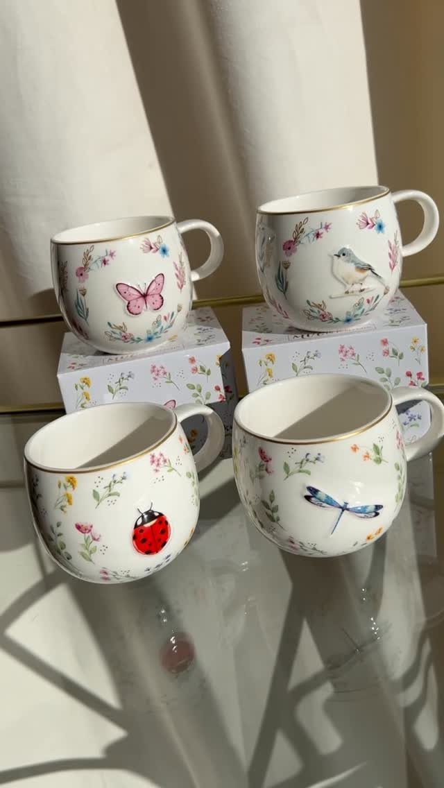 Four white ceramic mugs with nature illustrations and gold rims on a reflective surface.