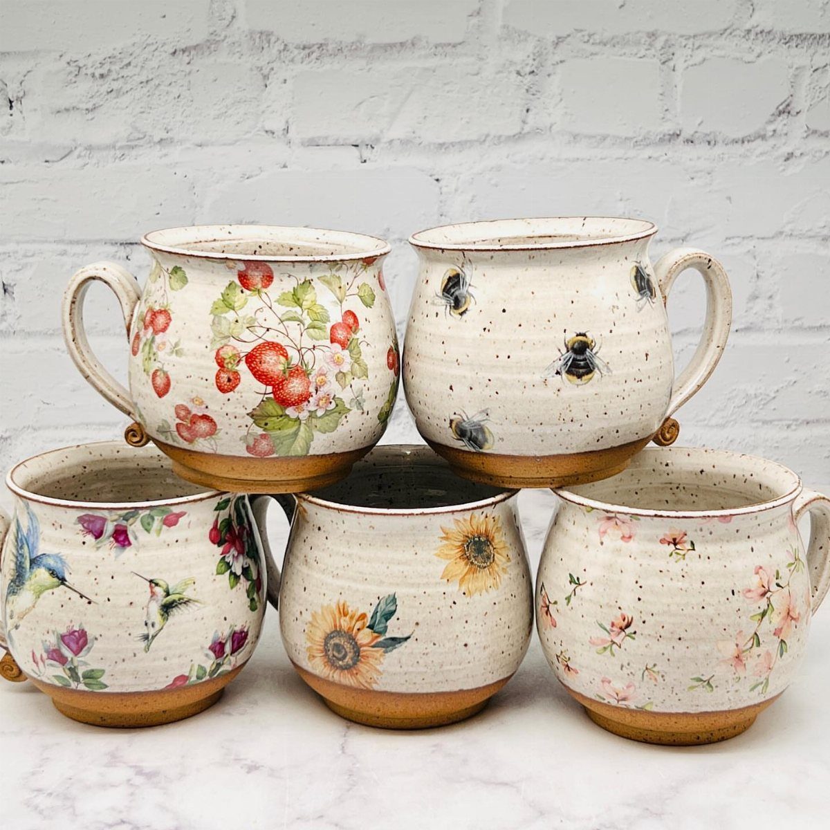 Five speckled stoneware mugs with floral and insect designs against a white brick wall.