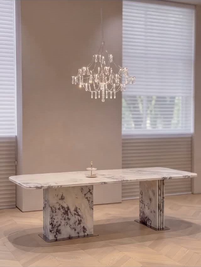 White marble dining table with pedestal legs under a crystal chandelier in a bright room.