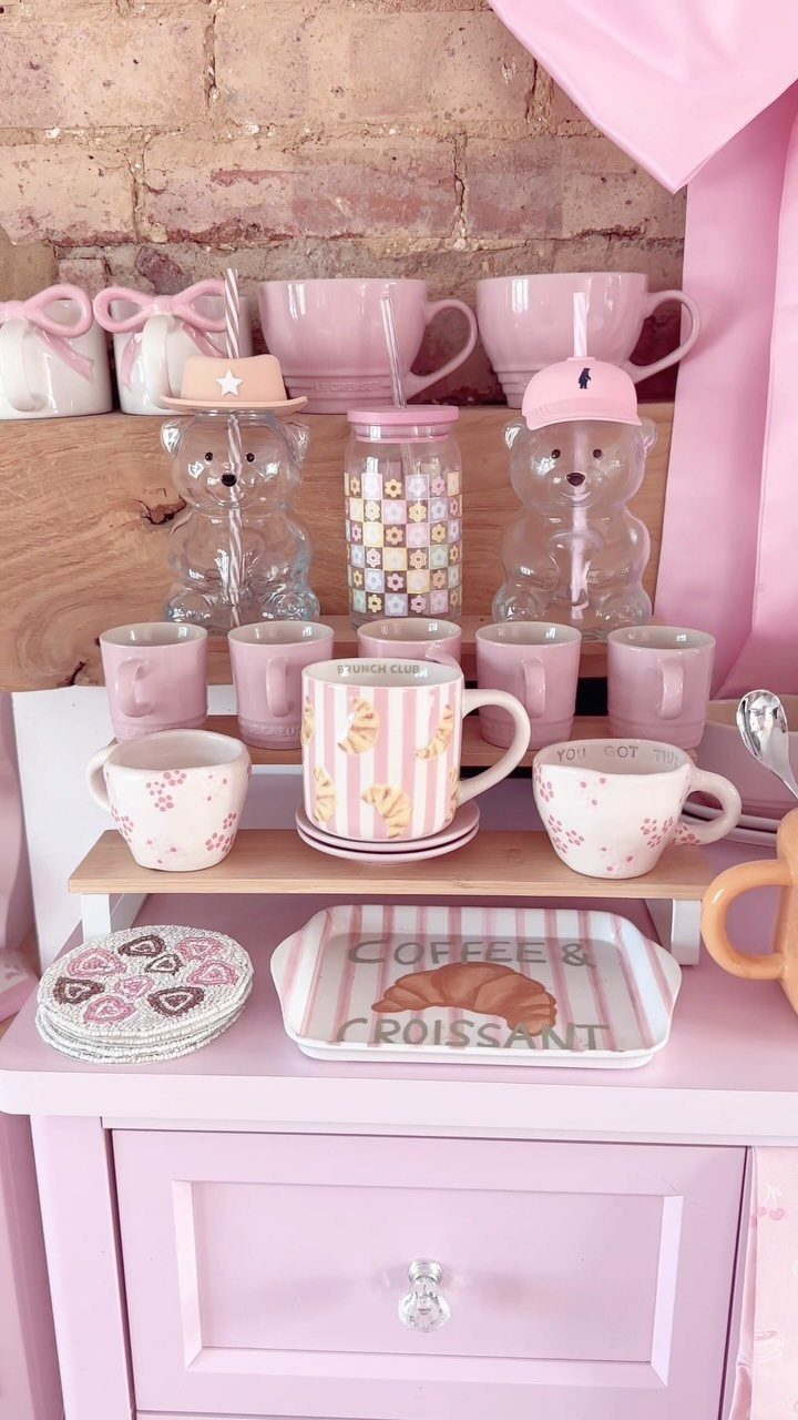 16 Home Coffee Stand Ideas to Fit Any Space 14 Pink coffee station with pink mugs, bear glasses, and a croissant tray on a cabinet.