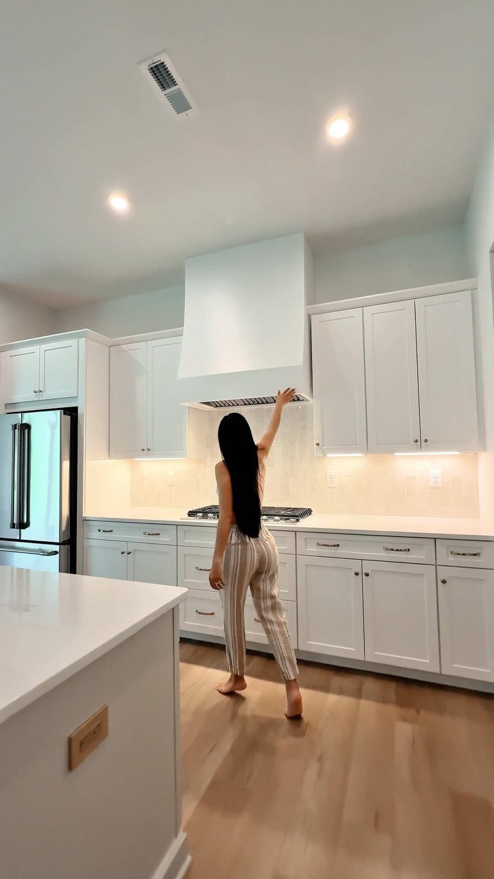 20 Home Kitchen Decor Ideas to Refresh Your Space 9 Woman standing in a white kitchen featuring a large minimalist decorative range hood.