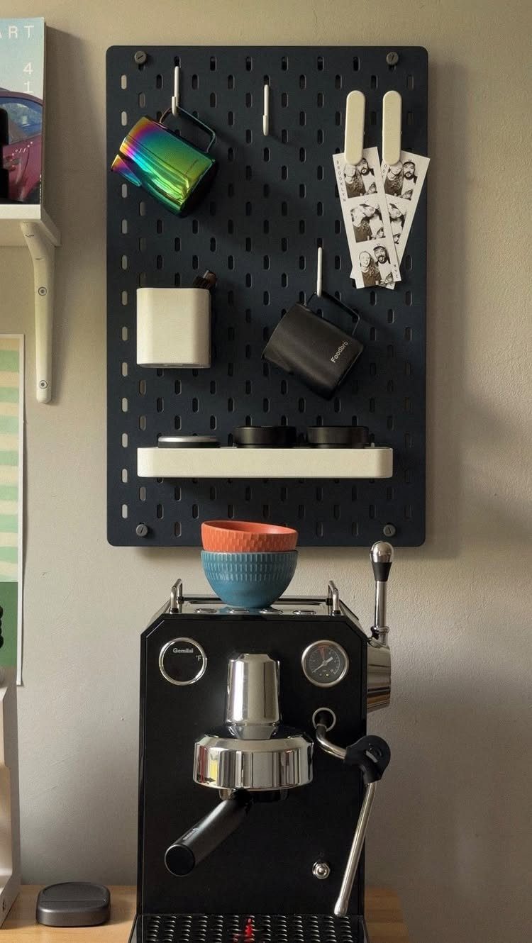 Black espresso machine and IKEA pegboard coffee station with mugs and accessories.