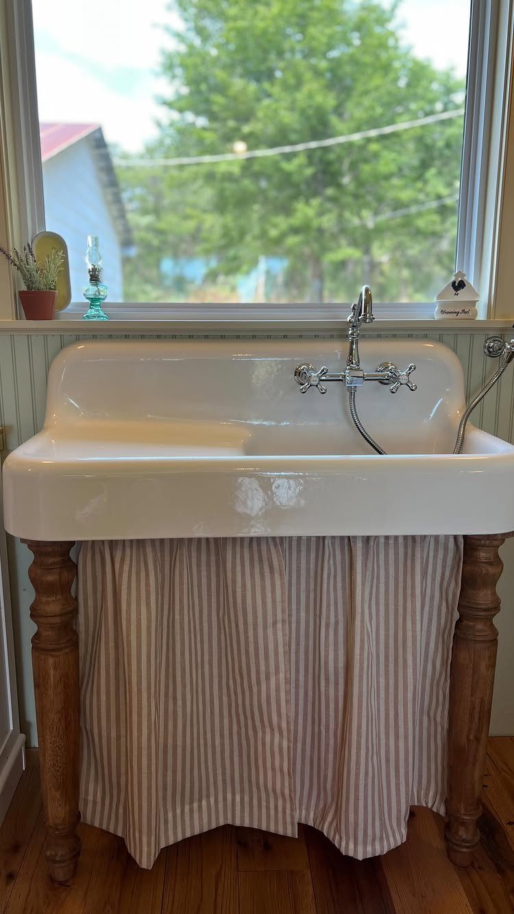 White apron-front farmhouse sink with wooden legs and a striped fabric curtain underneath.