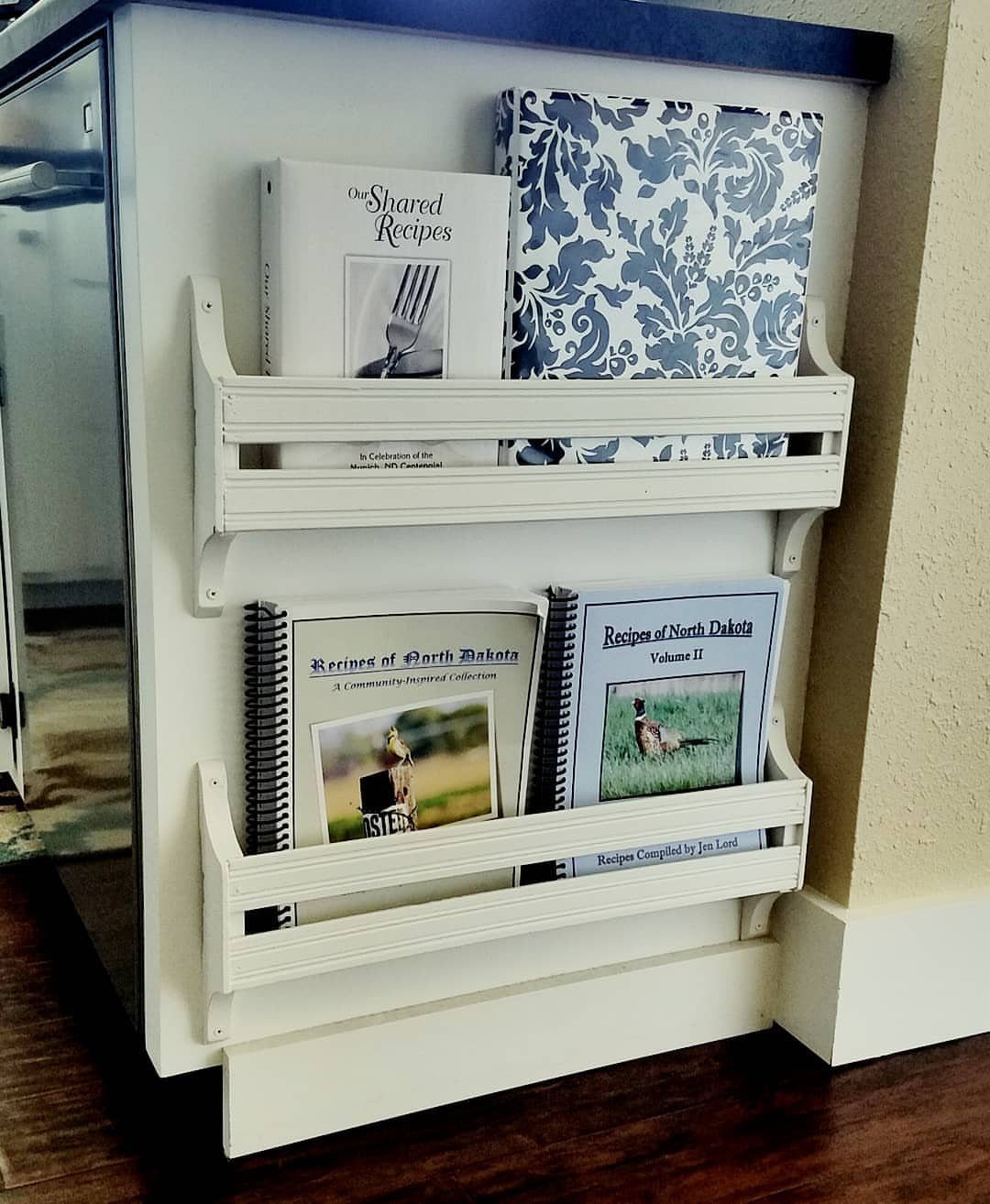 White wooden cookbook shelves mounted on the side of a white kitchen cabinet.