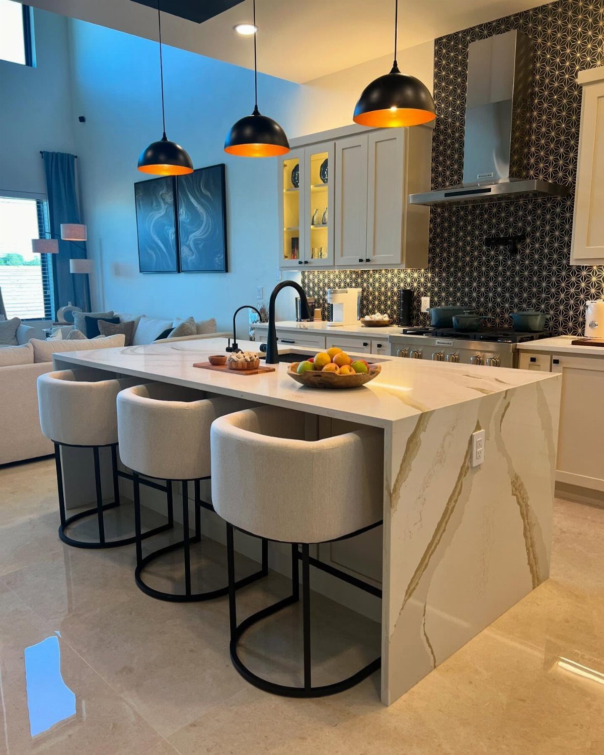 White marble waterfall kitchen island with three beige bar stools and black pendant lighting.