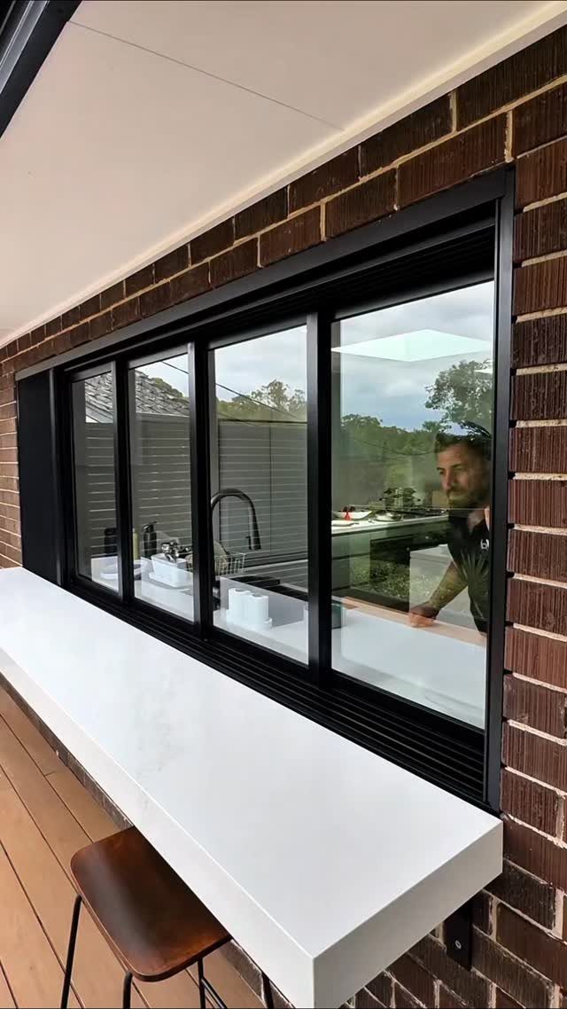 Black-framed sliding kitchen window with a white pass-through counter and outdoor stool.