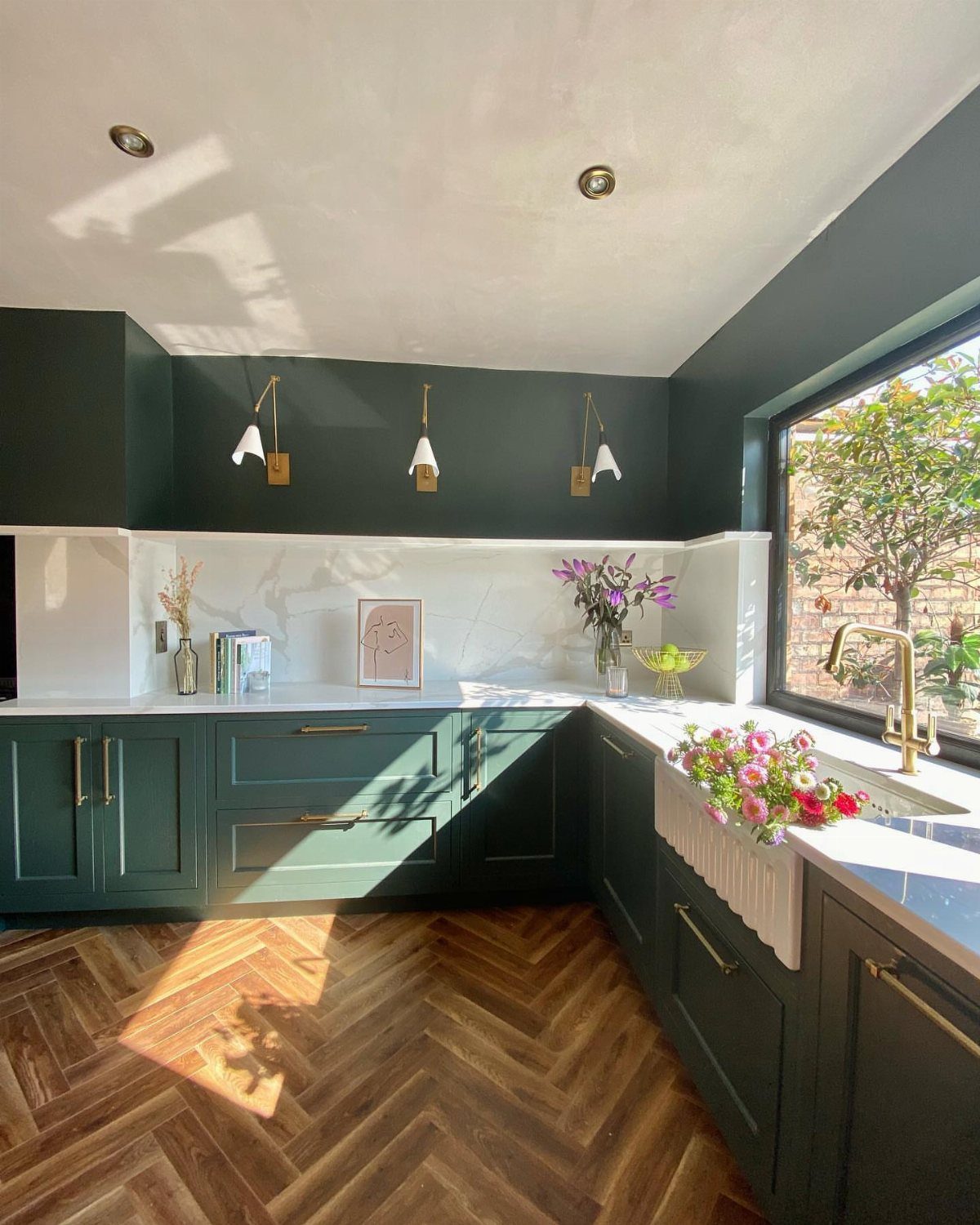 Kitchen window positioned above a white farmhouse sink with gold faucet and green cabinets.