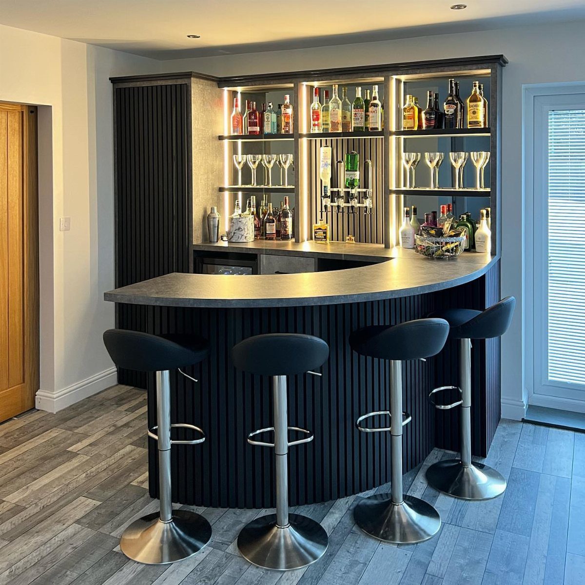 13 Mini Bar House Design Ideas to Fit Your Space and Taste 7 Curved modern home bar with black ribbed base, backlit shelves, and four bar stools.
