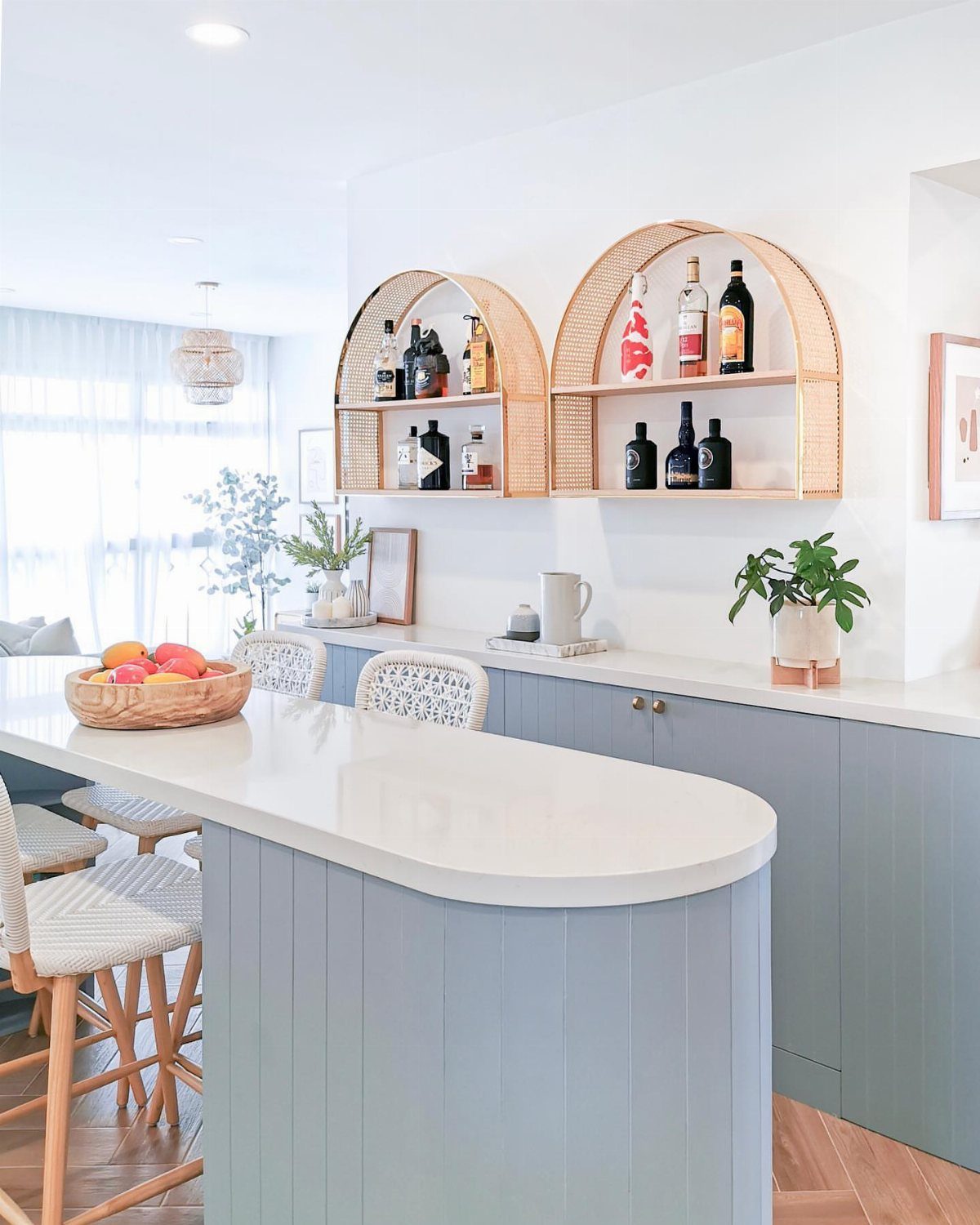 13 Mini Bar House Design Ideas to Fit Your Space and Taste 5 Light blue kitchen island with arched rattan wall shelves displaying liquor bottles and glassware.