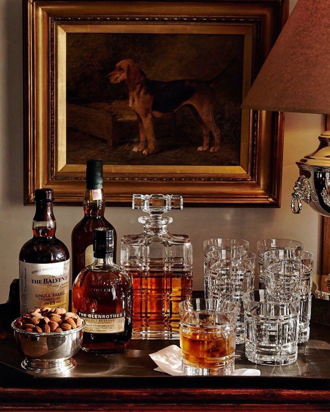 13 Mini Bar House Design Ideas to Fit Your Space and Taste 11 Liquor bottles and crystal glassware on a tray before a framed dog painting.