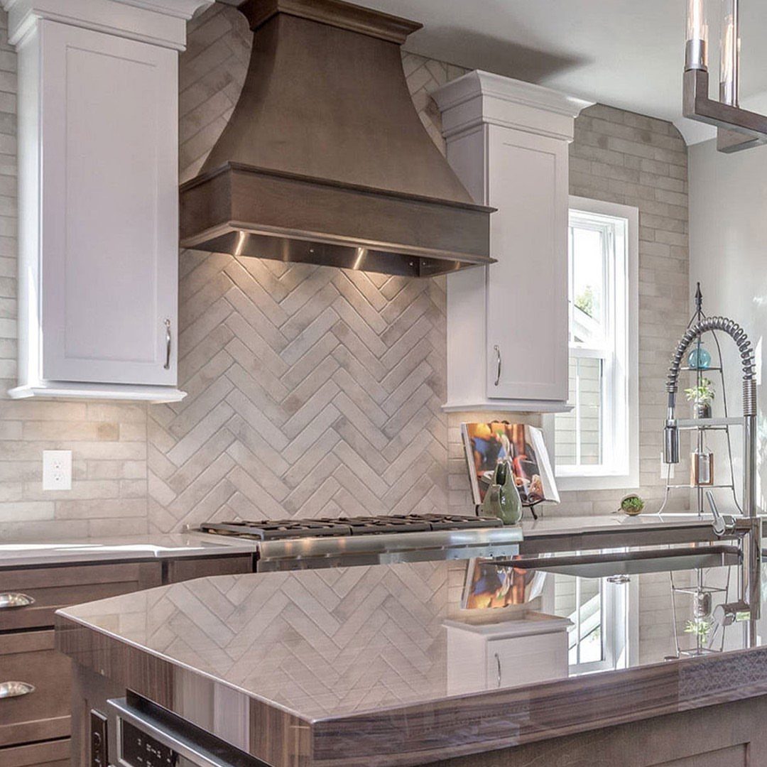 Herringbone pattern tile backsplash in a modern kitchen with white cabinets and metal hood.