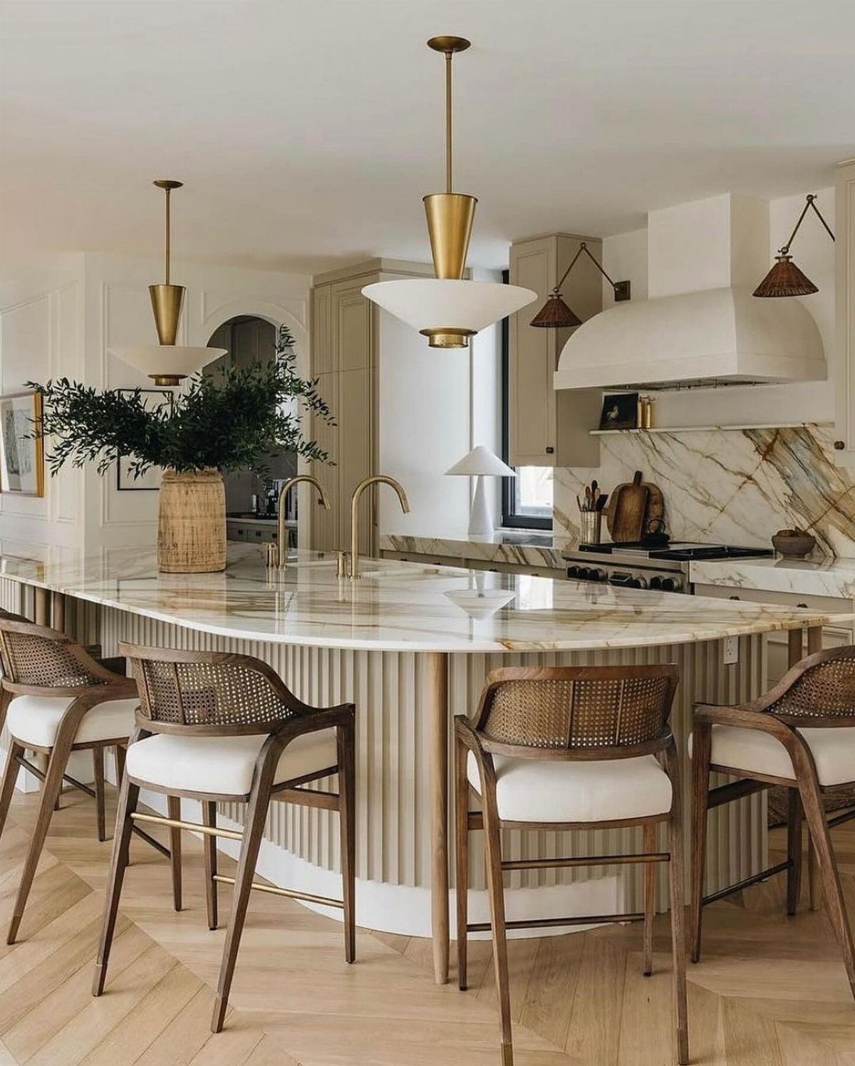 Curved white marble kitchen island with cane-back wooden stools and gold pendant lighting.