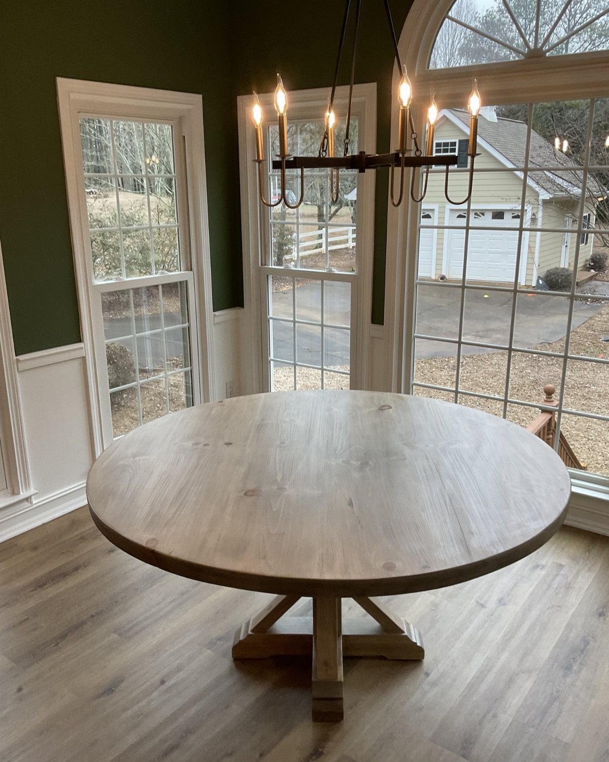 Round wooden pedestal table in a room with green walls and large windows.