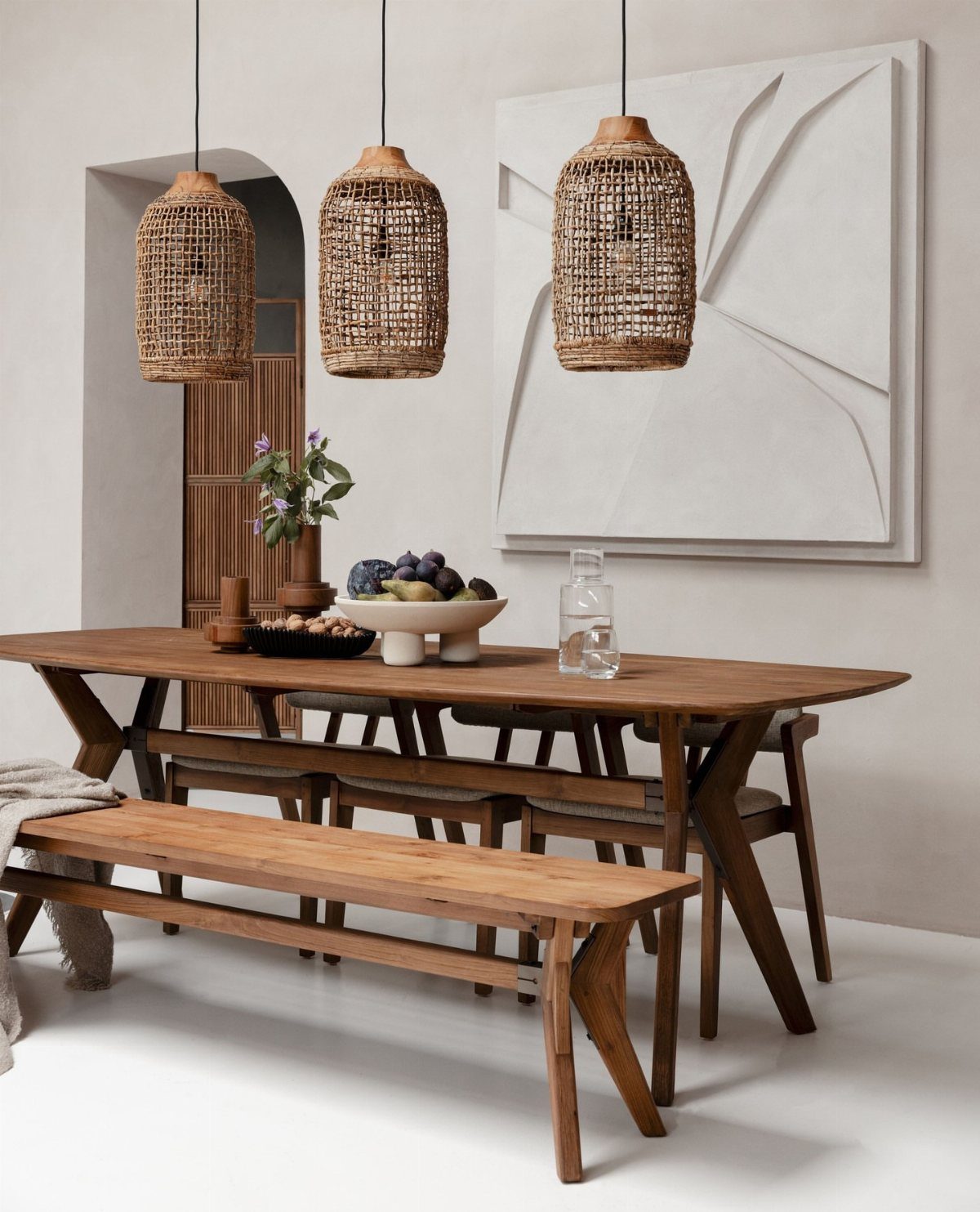 Wooden dining table with X-shaped legs and matching bench under woven wicker pendant lights.