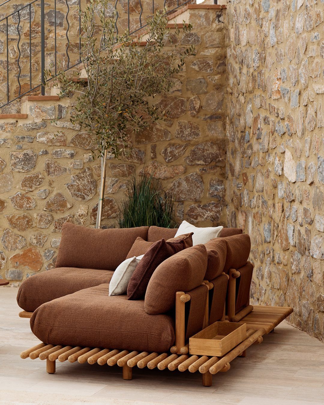 29 Scandinavian Outdoor Furniture for Patios and Gardens 29 Brown cushioned outdoor lounge sofa on a wooden base against a stone wall.