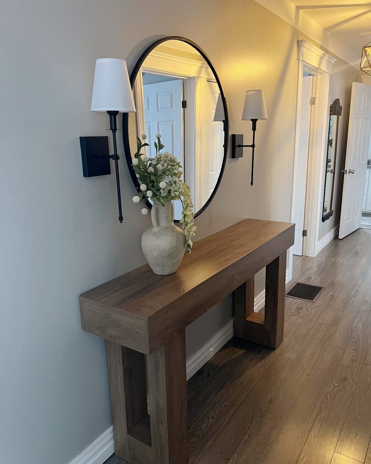 Wooden console table with a round mirror and wall sconces in a hallway.