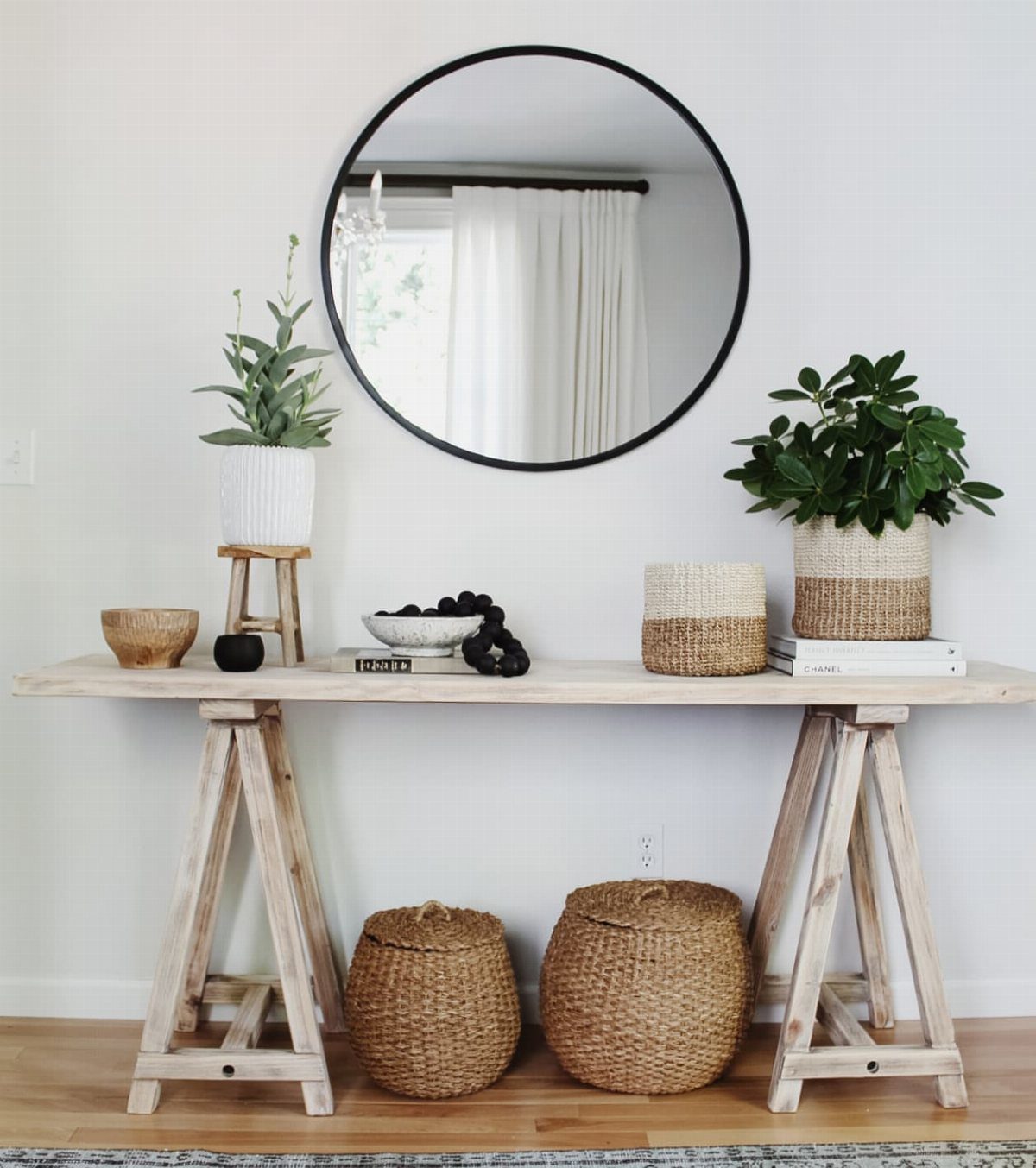 Light wood console table with potted green plants, woven baskets, and a round mirror.