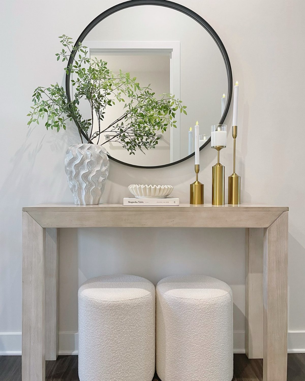 Light wood console table with green foliage, gold candles, and a round mirror.