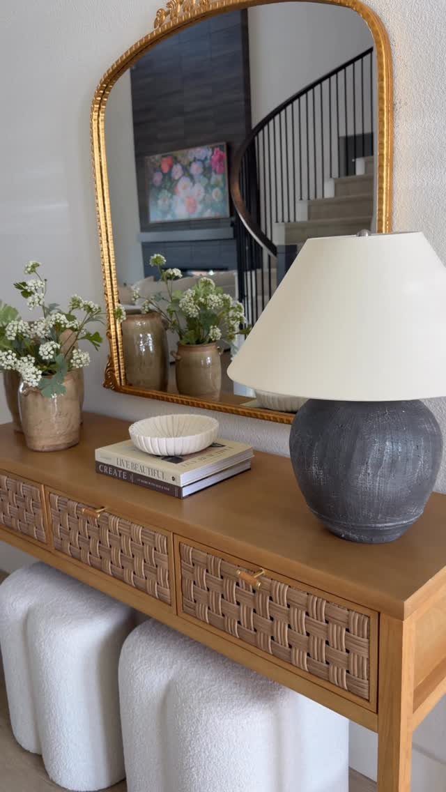 Wooden console table with a large grey lamp, white flowers, and gold mirror.