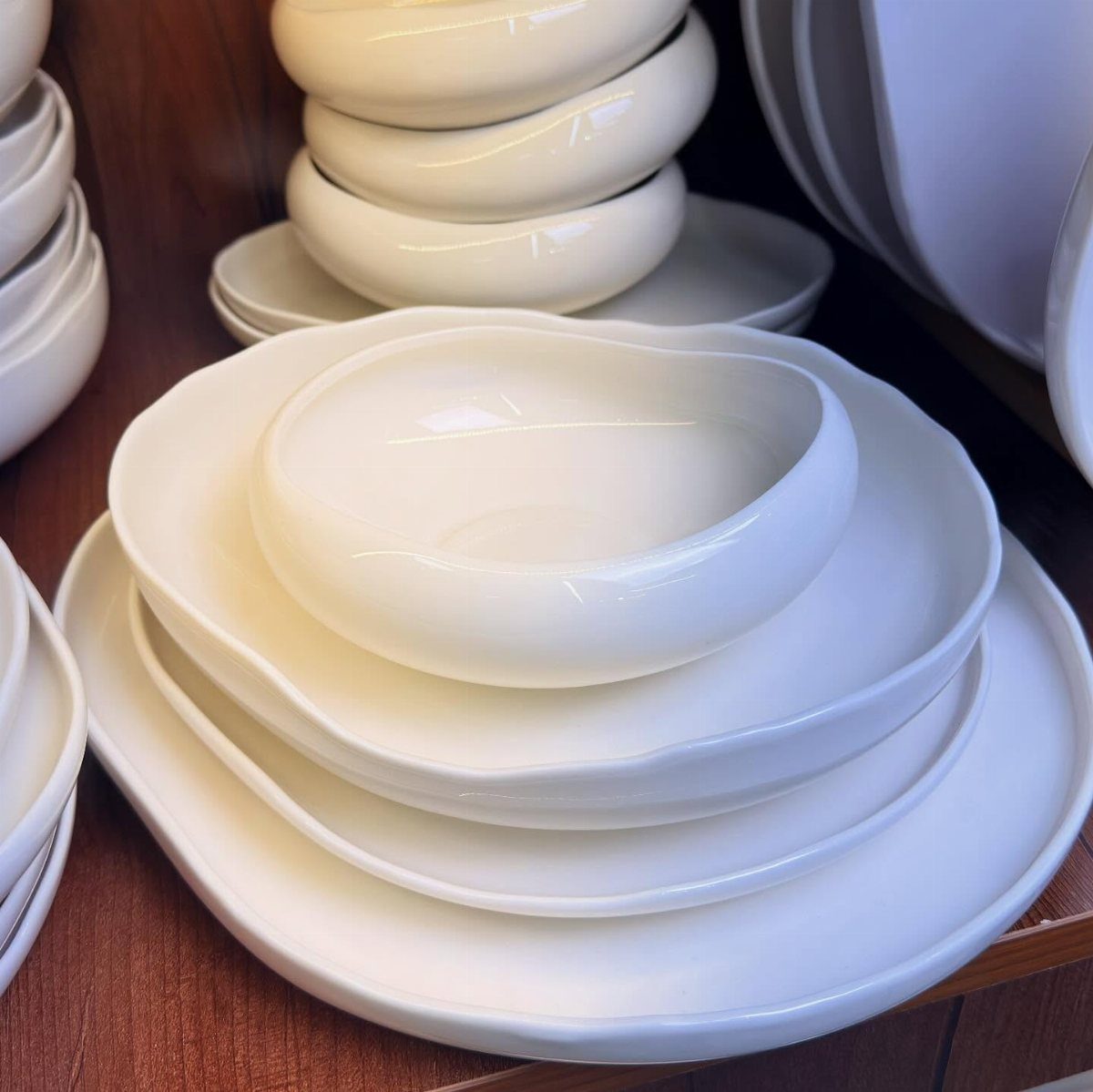 White glossy stoneware dishes with irregular organic shapes stacked on a wooden shelf.