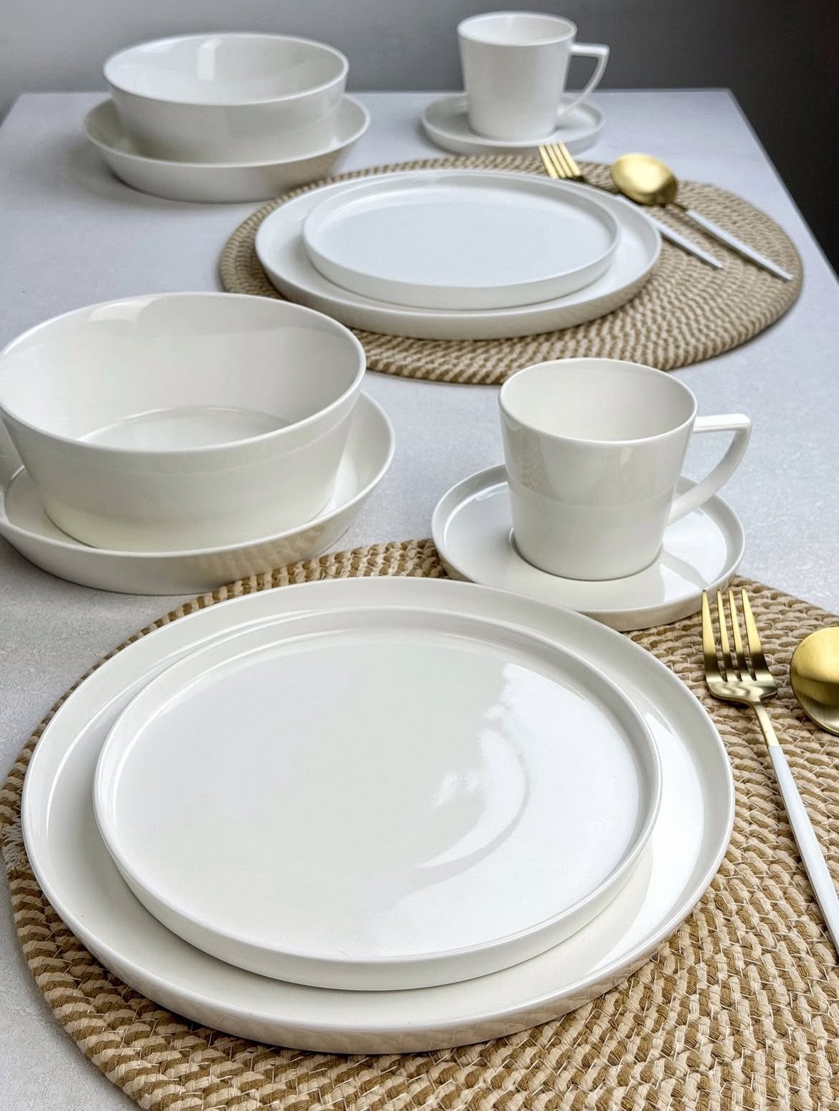 White rimmed stoneware plates, bowls, and mugs on woven placemats with gold cutlery.