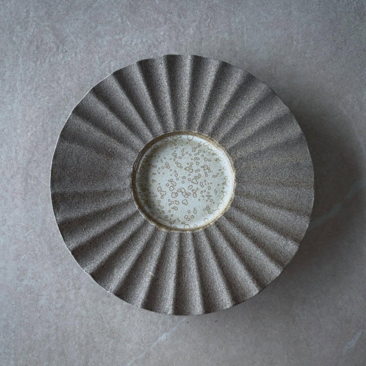 Grey stoneware plate with pleated texture and a speckled white center on stone.